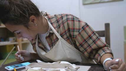 Young woman draws patterns on a clay product and looks at the sample in the phone. Pottery workshop.