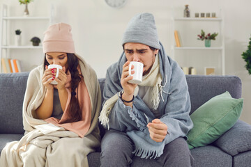 Sick young couple wrapped in plaids sitting on sofa, drinking hot tea and trying to warm up