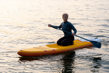 Fototapeta na wymiar A young athletic woman in a blue wetsuit floats across the sea on a yellow paddleboard holding an oar in the Golden rays of the dawn sun
