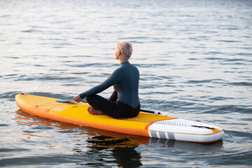 A young athletic woman in a blue wetsuit swims through the sea on a yellow paddle Board and meditates in the Golden rays of the dawn sun