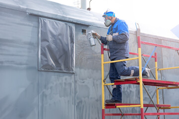 Industrial work. Priming of metal products from the compressor gun. A worker in overalls and a respirator paints the body of a truck trailer or a metal car.