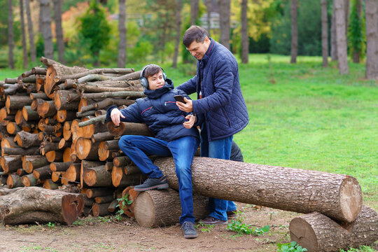 Teenager And Father Listening To Music By Headphones, Relaxing In Autumn City Park, Using Phone And Sitting On A Log, Beautiful Nature