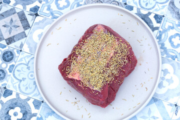 Raw beef roasting joint covered with rosemary on a grey plate.  Blue patterned tile background
