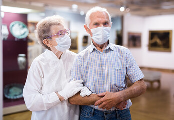 mature couple in mask protecting against covid examines paintings on display in hall of art museum