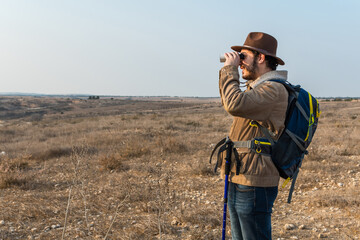 Hiker young man with beard, wearing brown jacket, backpack and a hipster hat look through binoculars.