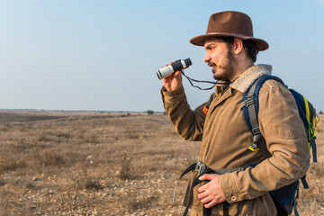 Fototapeta premium Hiker young man with beard, wearing brown jacket, backpack and a hipster hat look through binoculars.