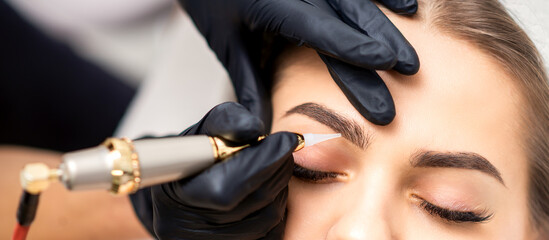 Beautician applying permanent makeup on eyebrows of young woman by special tattoo machine tool - obrazy, fototapety, plakaty