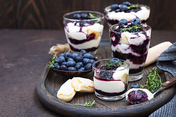 Dessert from mascarpone or ricotta cheese, blueberries and biscuit in glass on a dark background. No baked cheesecake or tiramisu. - obrazy, fototapety, plakaty