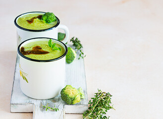 Two enamel mugs with healthy vegan broccoli soup with spicy oil and aromatic herbs over light background. Diet detox food concept. Copy space.