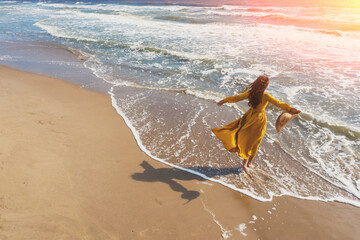 Fototapeta na wymiar Seascape on a summer sunny day. Woman on the beach. Young happy woman with hands in the air walks careless on the seaside in yellow fluttering dress. View from above