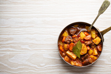 Delicious stew with meat, potatoes, carrot and gravy in rustic copper pot on white wooden...