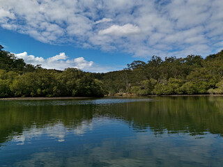 Obraz premium Beautiful morning view of a creek with reflections of deep blue sky, light clouds, mountains and trees, Cockle Creek, Bobbin Head, Ku-ring-gai Chase National Park, New South Wales, Australia 