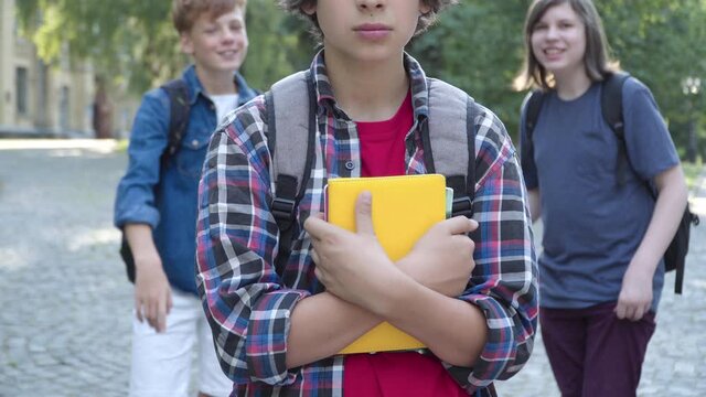 Unrecognizable nerd Caucasian boy standing with books outdoors as classmates laughing at him from the background. Bullied schoolboy suffering teasing on schoolyard.