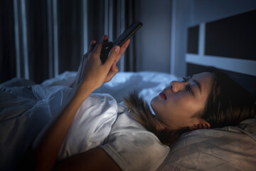 Beautiful Asian woman in bed using mobile smartphone technology, tired addictive can’t sleep,...