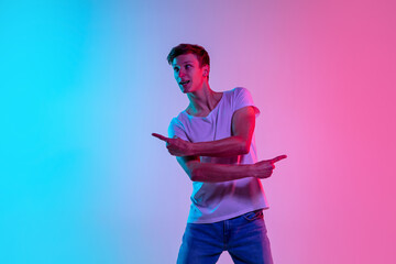 Pointing at sides. Young caucasian man's portrait on gradient blue-pink studio background in neon light. Concept of youth, human emotions, facial expression, sales, ad. Beautiful model in casual.