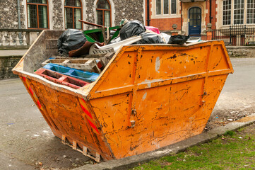 Heavy metal orange iron skip container full of scrap waste household rubbish such as black plastic bin bags and pallets all destined for a landfill site, which causes environmental pollution