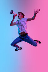 Fototapeta na wymiar Shouting with loudspeaker. Young man's jumping high on gradient blue-pink studio background in neon light. Concept of youth, human emotions, facial expression, sales, ad. Beautiful model in casual.