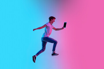 Fototapeta na wymiar Winner with tablet. Young caucasian man's jumping high on gradient blue-pink studio background in neon light. Concept of youth, human emotions, facial expression, sales, ad. Beautiful model in casual.