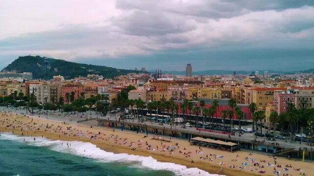 Aerial view of people at the Playa de Barceloneta Beach and the city of Barcelona, gloomy evening, in Spain - pull back, drone shot