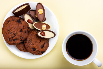 Chocolates, a cup of coffee and cookies with chocolate chips on a yellow background. View from above.