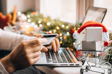 Christmas online shopping. male buy order on laptop computer xmas presents. man using credit bank card at home lockdown. gift boxes in trolley. Winter New Year holidays sales, discounts, promotions.