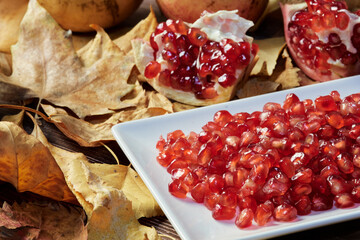 closeup of red seeds of the pomegranate fruit prepared on a plate ready to eat and a split pomegranate in the background. background of dry fallen leaves of trees in autumn