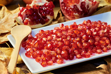 close-up of red seeds of the pomegranate fruit prepared on a plate ready to eat with a wooden spoon. background of dry fallen leaves of trees in autumn