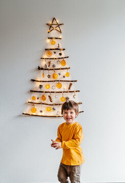 Little child playing with handmade craft Christmas tree made from sticks and natural materials hanging on wall. Sustainable Christmas, zero waste, plastic free, eco friendly.