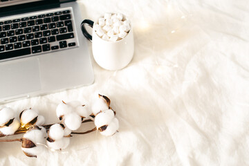 Laptop and cup of cocoa with marshmallows, cotton flowers, sparkle lights on white bed. Work at home concept. Autumn, fall, winter composition. Flat lay, top view, copy space.