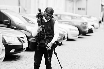 Young professional african american videographer holding professional camera with tripod pro equipment. Afro cameraman wearing black duraq making a videos.