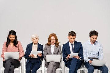 Multicultural businesspeople using devices while sitting on chairs isolated on grey