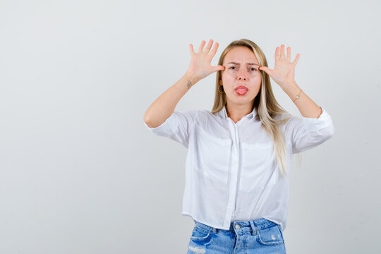  Blonde Lady In Blouse, Shorts Holding Hands Near Head As Ears, Sticking Out Tongue And Looking Grumpy , Front View.