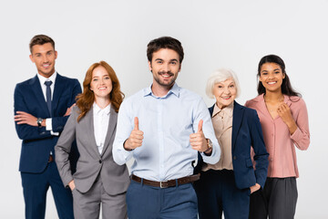 Smiling businessman showing thumbs up near cheerful multiethnic colleagues isolated on grey