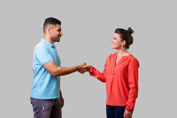 Portrait of positive friendly man and woman in casual wear standing, smiling at each other and shaking hands, couple first meeting or acquaintance. isolated on gray background, indoor studio shot