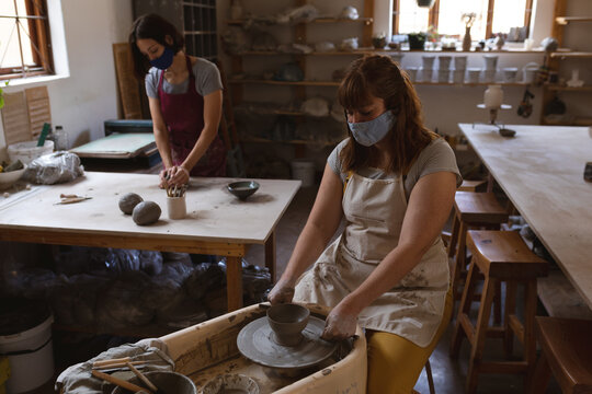Two caucasian female potters in face masks working in pottery studio