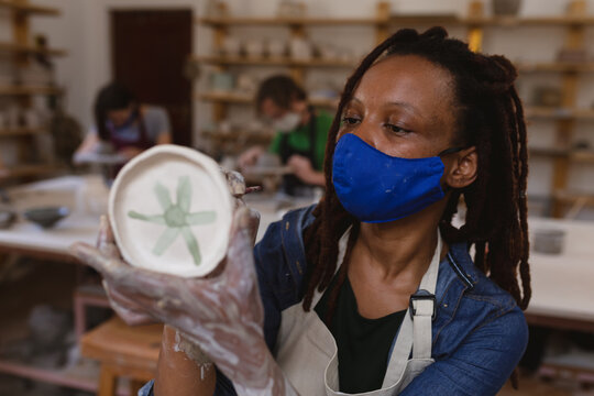 Mixed race female potter in face mask working in pottery studio - Powered by Adobe