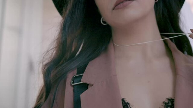 Close up of fashionable brunette young confident woman wearing a pink coat and a black hat playing with her necklace. Slow motion, 4k, parallax, cinematic, sun flare