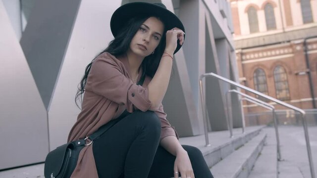 Close up of fashionable brunette young confident woman wearing a pink coat and black hat sitting on the stairs and looking into the camera dramatic and feminine. Slow motion, 4k, frontal view, push in