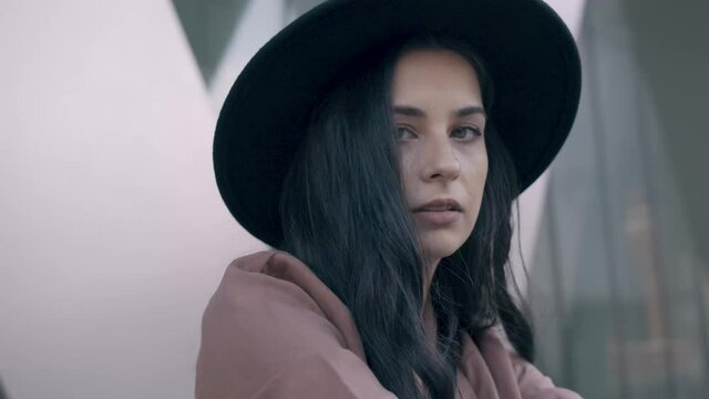 Close up of fashionable brunette young confident woman wearing a pink coat and black hat sitting on the stairs and looking into the camera. Slow motion, 4k, frontal view, parallax, cinematic