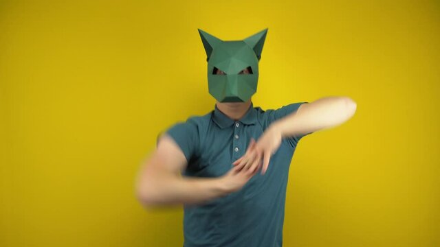 Young man in a cardboard jackal mask dances on a yellow background. Man in a green polo and mask.