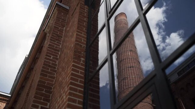 Old chimney of Moscow Arma business quarter reflected in a window. Action. Bottom view of a red brick wall of an old gas factory with a high chimney on blue cloudy sky background.