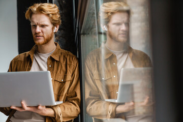 Brooding redhead guy in wireless earphones working with laptop