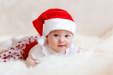 Cute funny five-month-old baby girl in a white bodysuit and red santa cap lies on her stomach on fluffy plaid. newborn babies for the new year