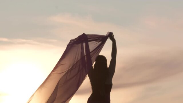 A beautiful woman in an oriental costume walks on the sand at sunset. Raises a transparent veil overhead that flutters in the wind. Shooting from behind.   Silhouette.