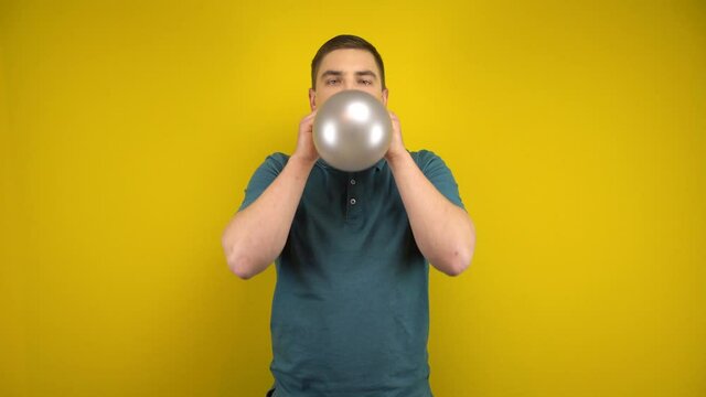 A young man inflates a silver balloon with his mouth on a yellow background. Man in a green polo.