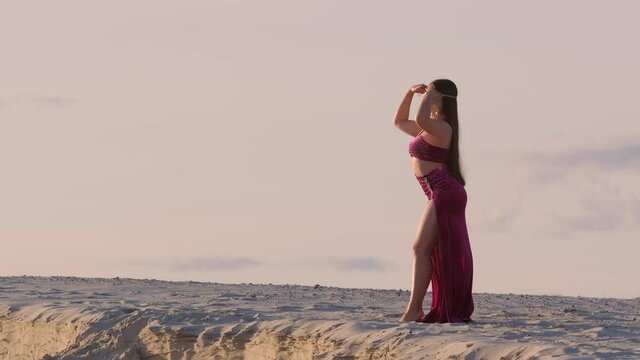 Beautiful young woman in oriental costume with jewelry on her head in oriental style dancing belly dance on sand against the sky at sunset.