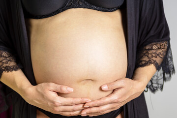 Eine schwangere Frau bekleidet mit schwarzer Unterwäsche hält ihren Babybauch mit ihren beiden Händen - obrazy, fototapety, plakaty
