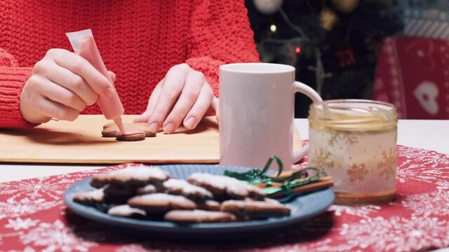 A young beautiful woman paints ginger cookies with pink cream. There is a Cup of cocoa on the table. Cooking Christmas gingerbread ginger cookies.