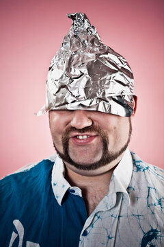 Studio photo of a goofy looking man wearing a tin foil hat on dark pink background.