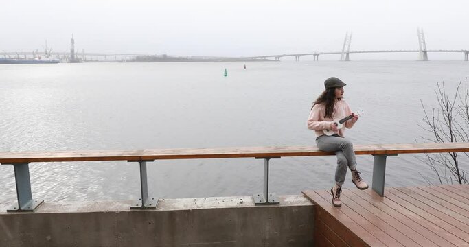 An attractive curly-haired girl sits on wooden fence of city seafront on a cloudy day, playing a white musical instrument ukulele and singing songs